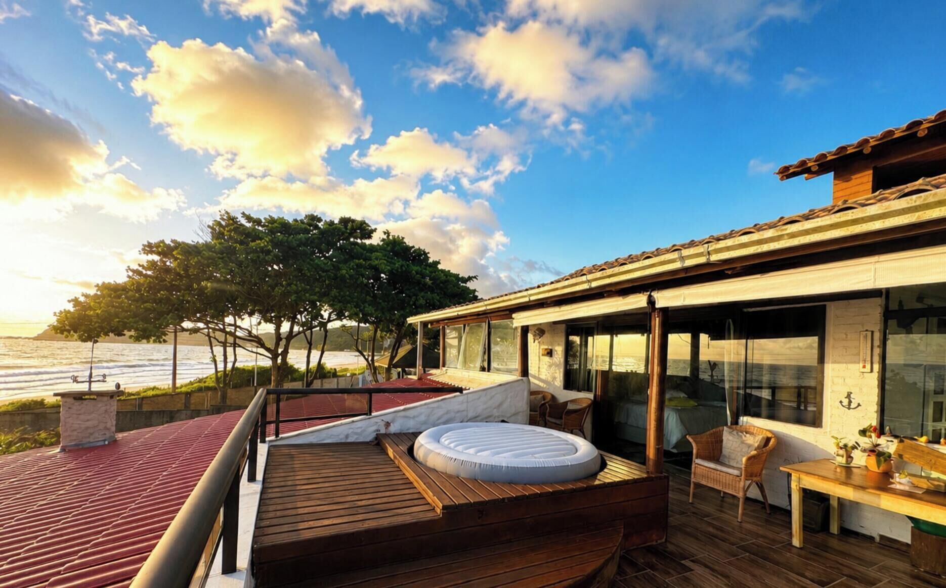 Una terraza de madera con un espacio de estar circular, una habitación con paredes de vidrio y vistas al mar, y un techo de tejas rojas.