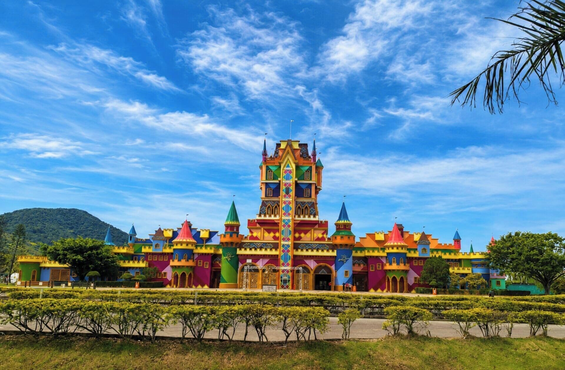 A colorful castle-like structure with multiple towers and a central spire, surrounded by well-maintained greenery.