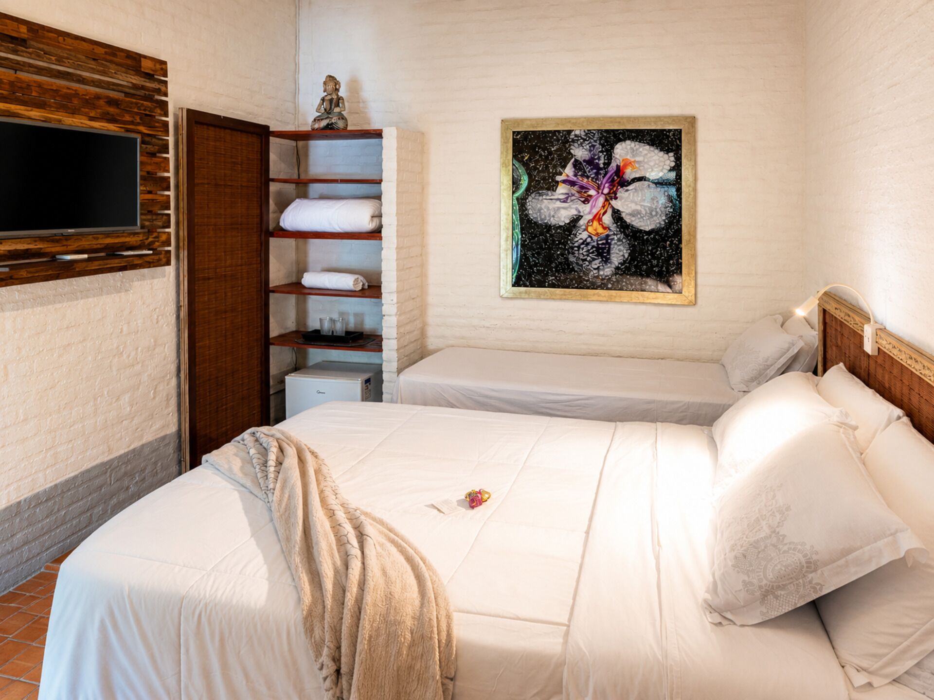 A hotel room with two beds, a flat-screen TV mounted on the wall, a shelf with folded towels, and a painting on the wall.