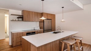 Ett modernt kök med träskåp, en vit bänkskiva, en inbyggd diskho och ett kylskåp i rostfritt stål. Det finns två barstolar och hänglampor ovanför bänkskivan.
