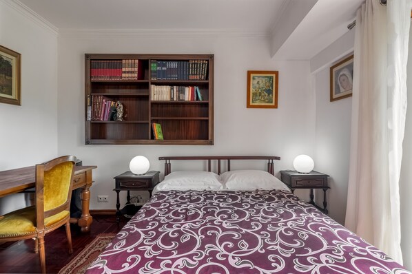 A bedroom with a bed, a wooden bookshelf, a desk, a chair, and two bedside tables with lamps.