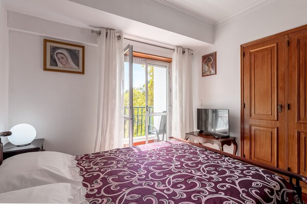Un dormitorio con cama, televisor, armario y balcón con cortinas.