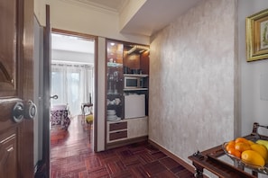 Una habitación con puerta de madera, una puerta de cristal que da a otra habitación, un refrigerador pequeño, un microondas y un frutero sobre una mesa de madera.