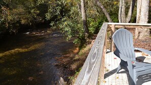 A wooden deck with a chair overlooking a river, surrounded by trees. - Super Cute River Front tiny home in Lake Toxaway. (Lake Toxaway)