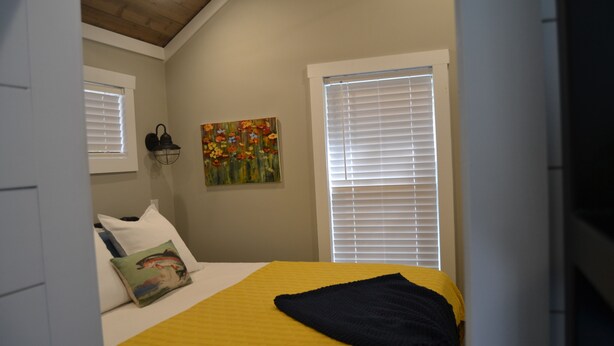 A bedroom with a bed, a painting, a wall-mounted lamp, and a window with blinds. - Super Cute River Front tiny home in Lake Toxaway. (Lake Toxaway)