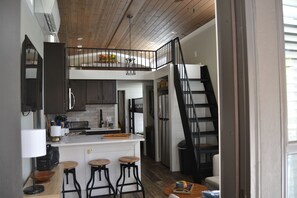 A modern kitchen with a central island, bar stools, a loft bedroom above, and a staircase.