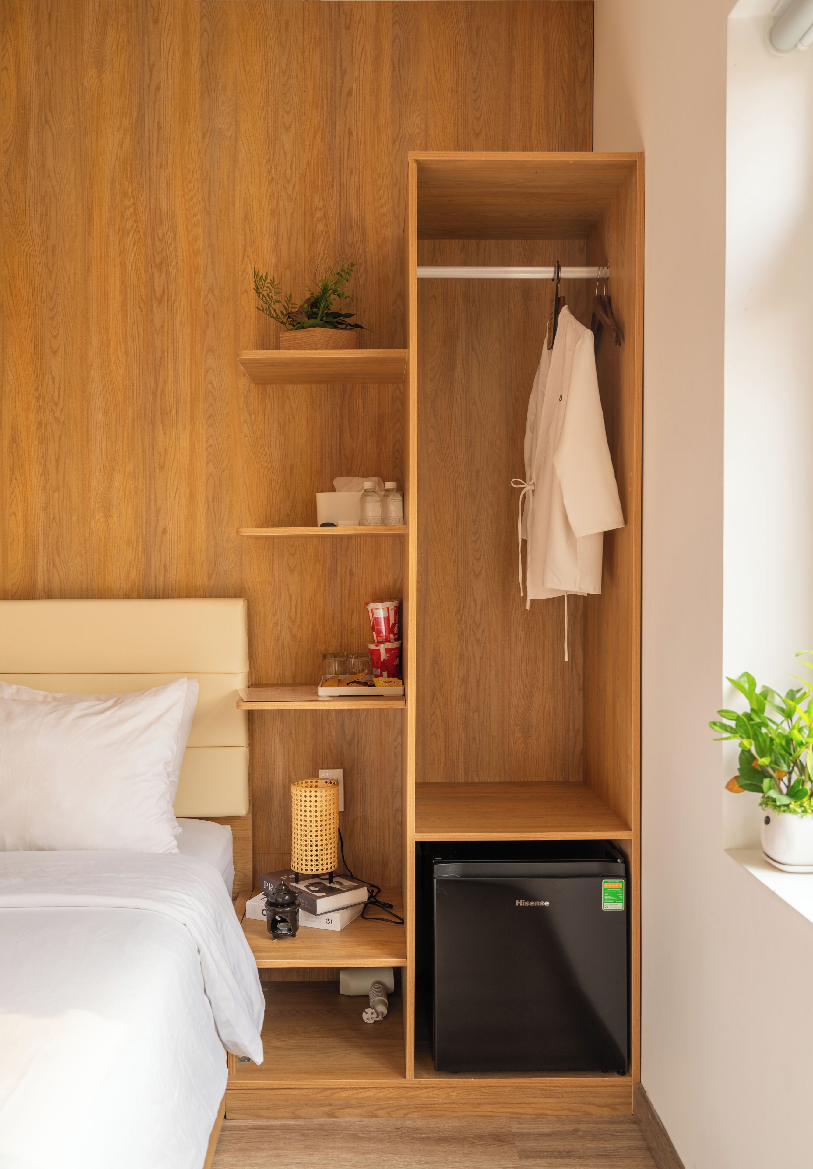 A compact room with a bed, a wooden wardrobe, a small black refrigerator, and a shelf with a plant.