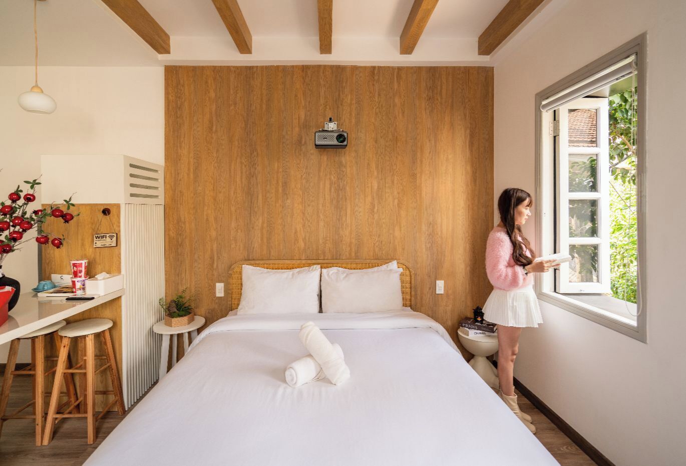 A hotel room with a wooden headboard, a bed with white linens, a desk with a computer, and a window with a view of greenery.