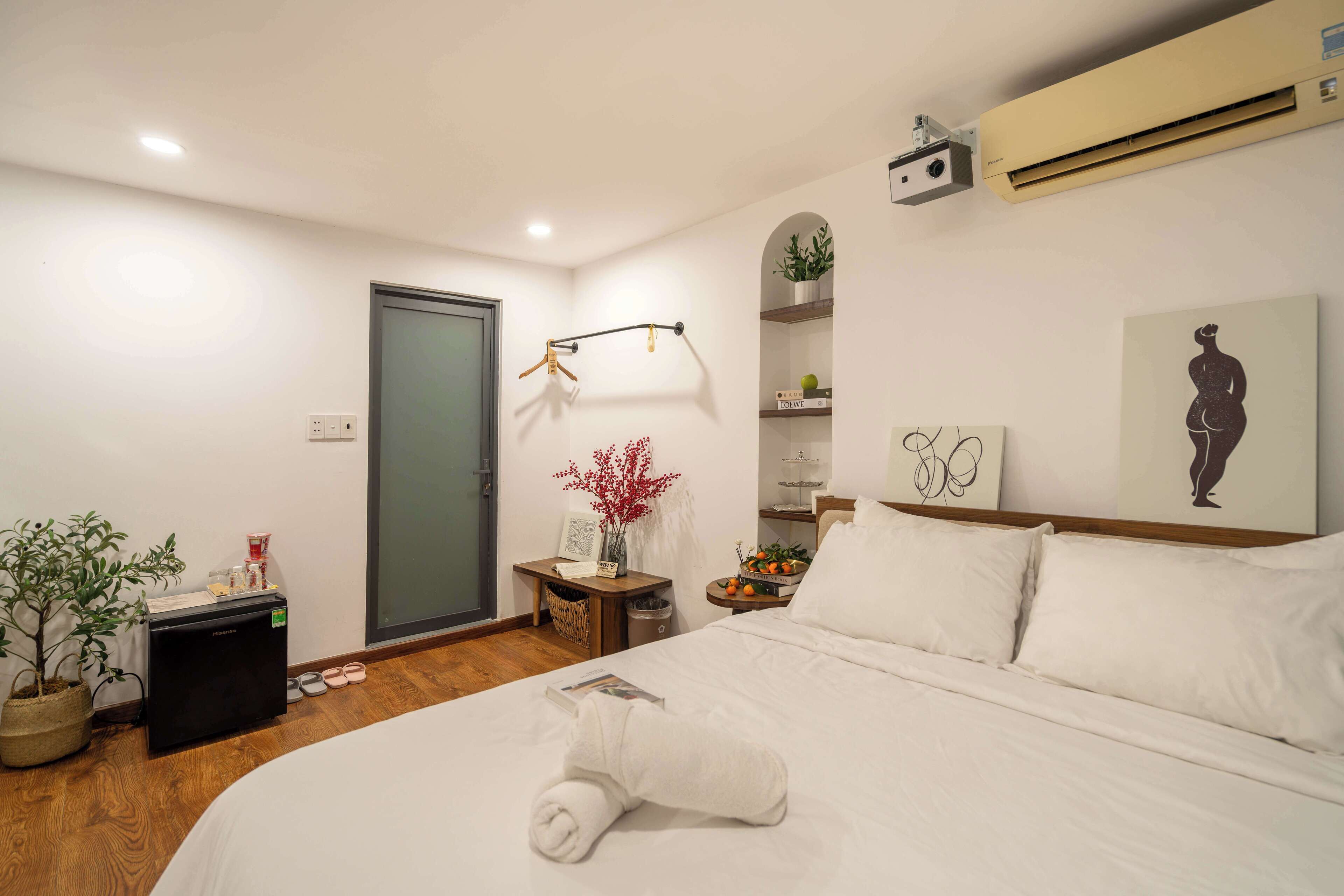A neatly arranged hotel room with a bed, a small desk, a potted plant, a wall-mounted air conditioner, and a door.