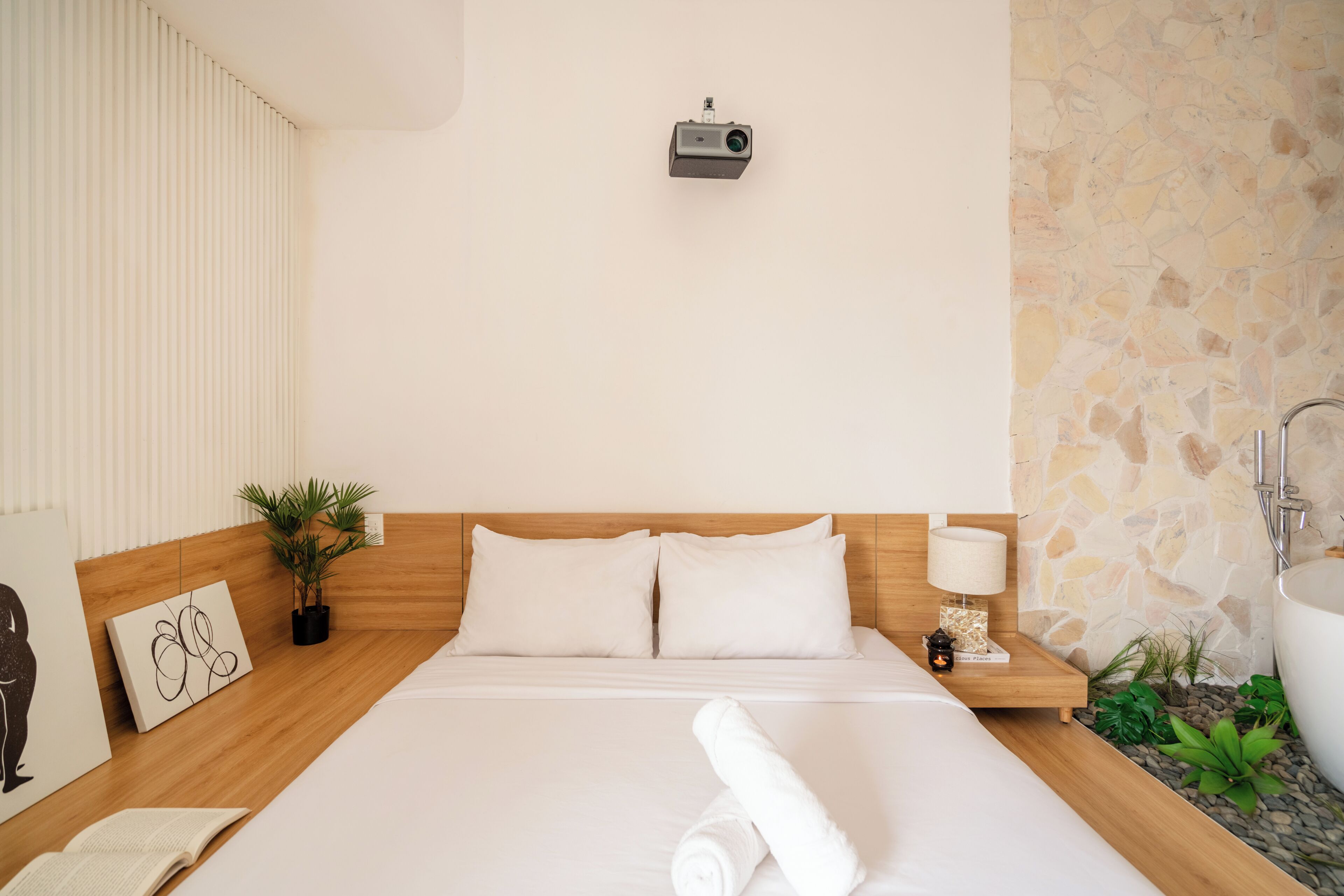 A hotel room with a bed, a nightstand with a lamp, a wall-mounted projector, and a stone wall.