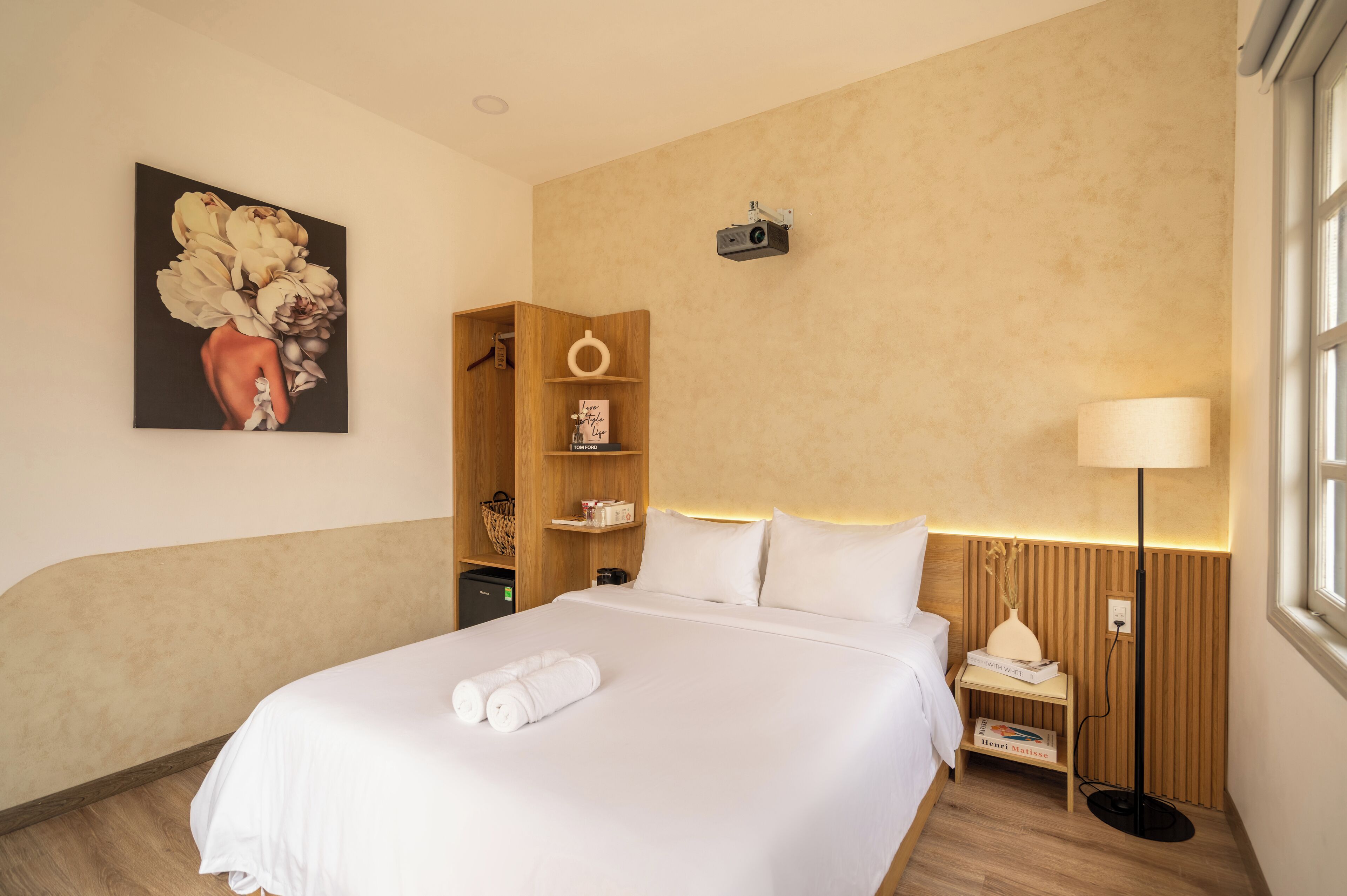 A hotel room with a large bed, a wooden wardrobe, a wall-mounted projector, and a painting of flowers.