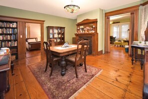 A dining room with a wooden table and chairs, a fireplace, a bookshelf, and a stained glass ceiling light. - Balcony Room in Victorian Home private bath (Shelburne Falls)
