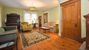 A living room with a piano, a green armchair, a patterned rug, a wooden door, and a chandelier. - Balcony Room in Victorian Home private bath (Shelburne Falls)