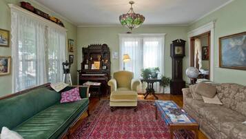 Een gezellige woonkamer met een groene bank, een beige fauteuil, een kamerkleed met patroon, een piano en een groot schilderij aan de muur.