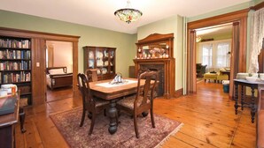 Uma sala de jantar com mesa de madeira, cadeiras, lareira, estante de livros e luminária no teto em vitral.