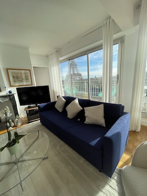 A modern living room with a large window, a flat-screen TV, a blue sofa, and a glass coffee table.