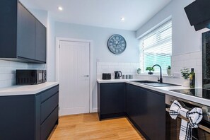A modern kitchen with dark cabinetry, a white countertop, a black clock, and a window with blinds.