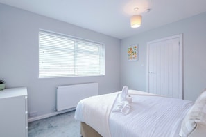 A bedroom with a bed, a radiator, a window with blinds, a door, and a wall-mounted light fixture.