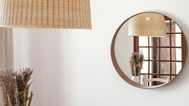 Un coin repas avec une table en bois, des chaises, un luminaire suspendu en fibres tressées, un miroir rond et un vase contenant des plantes séchées.