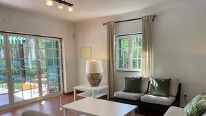 Un salon avec un canapé blanc, une table blanche, une lampe et une vue sur l’extérieur grâce à une porte vitrée.