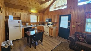 A wooden cabin with a kitchen featuring a refrigerator, stove, microwave, and wooden cabinets. There is a dining table with chairs and a couch in the living area.