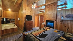 A wooden cabin interior with a large bed, a television, a fireplace, and a hot tub.
