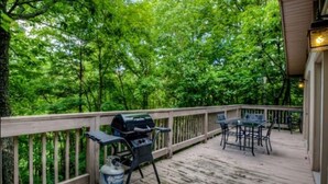 A wooden deck with a grill and table, surrounded by lush greenery.