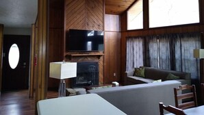 Una sala de estar acogedora con chimenea, un televisor de pantalla plana montado en una pared de madera, un sofá y una mesa de comedor con sillas.