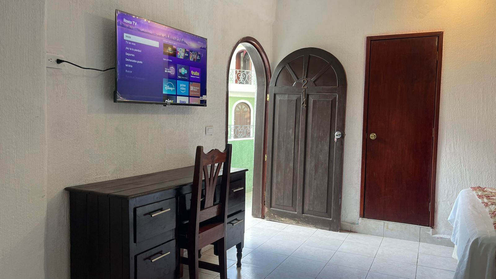 A room with a flat-screen TV mounted on the wall displaying a streaming service interface, a wooden desk with drawers, a chair, and a large wooden door with a window.