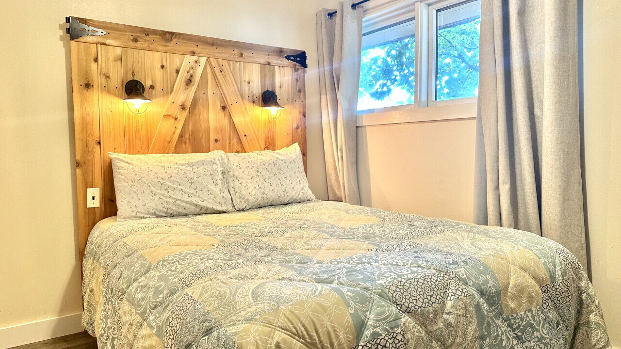 A bed with a patterned bedspread, a wooden headboard with two wall-mounted lights, and a window with curtains.