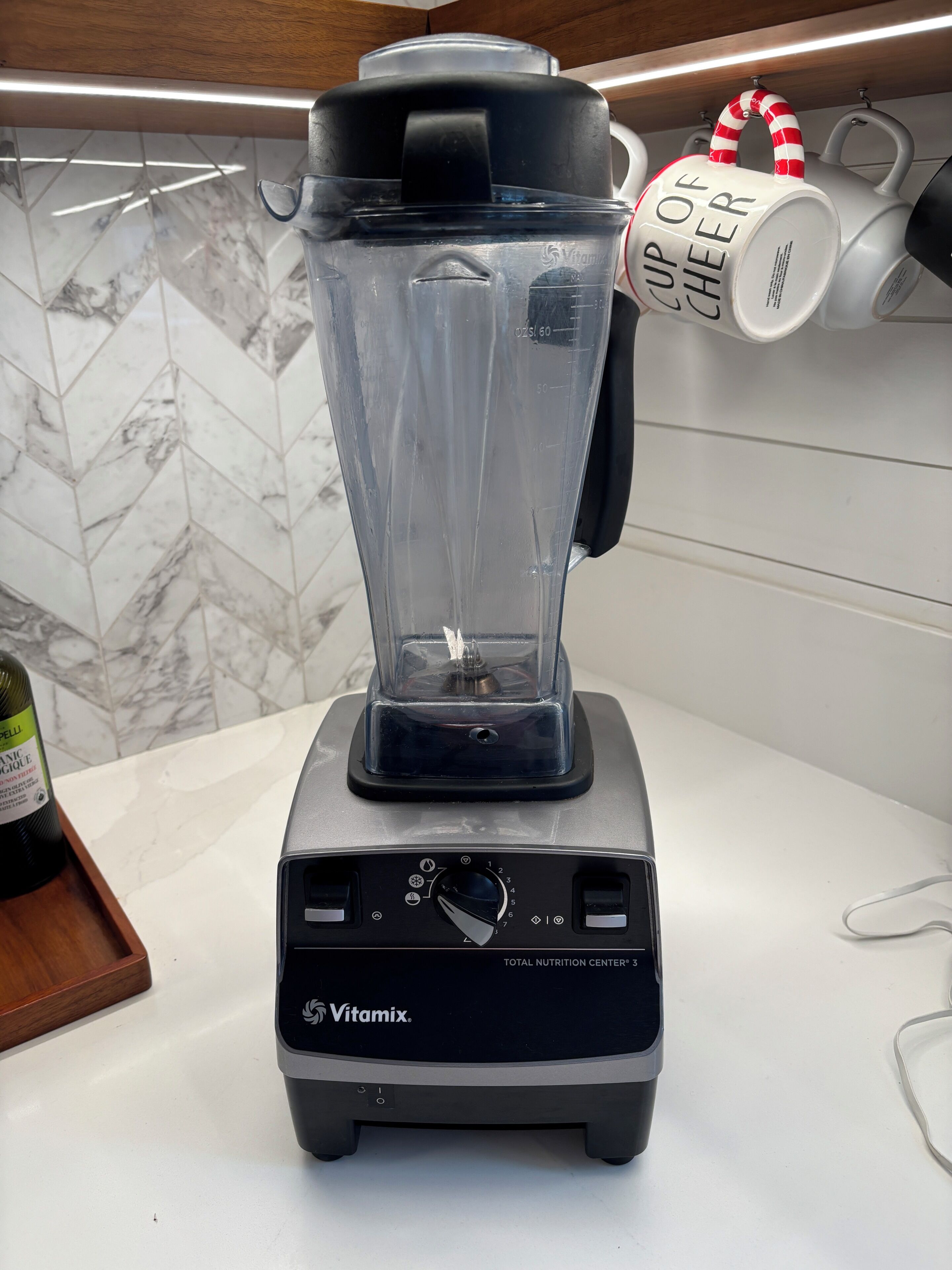 Een Vitamix blender op een aanrecht met een marmeren achterwand en een mok met de tekst 'Cup of Cheer'.