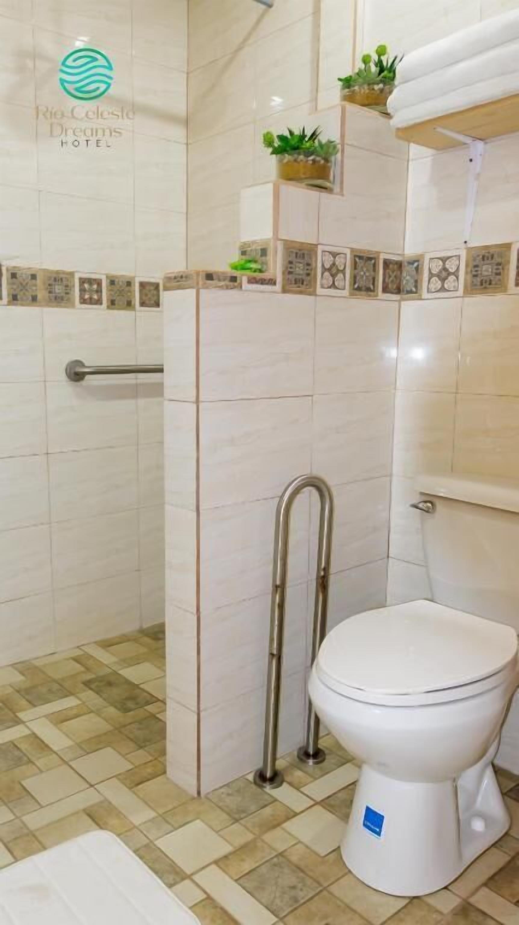 Un baño con inodoro, ducha con barra de apoyo y piso de baldosas.