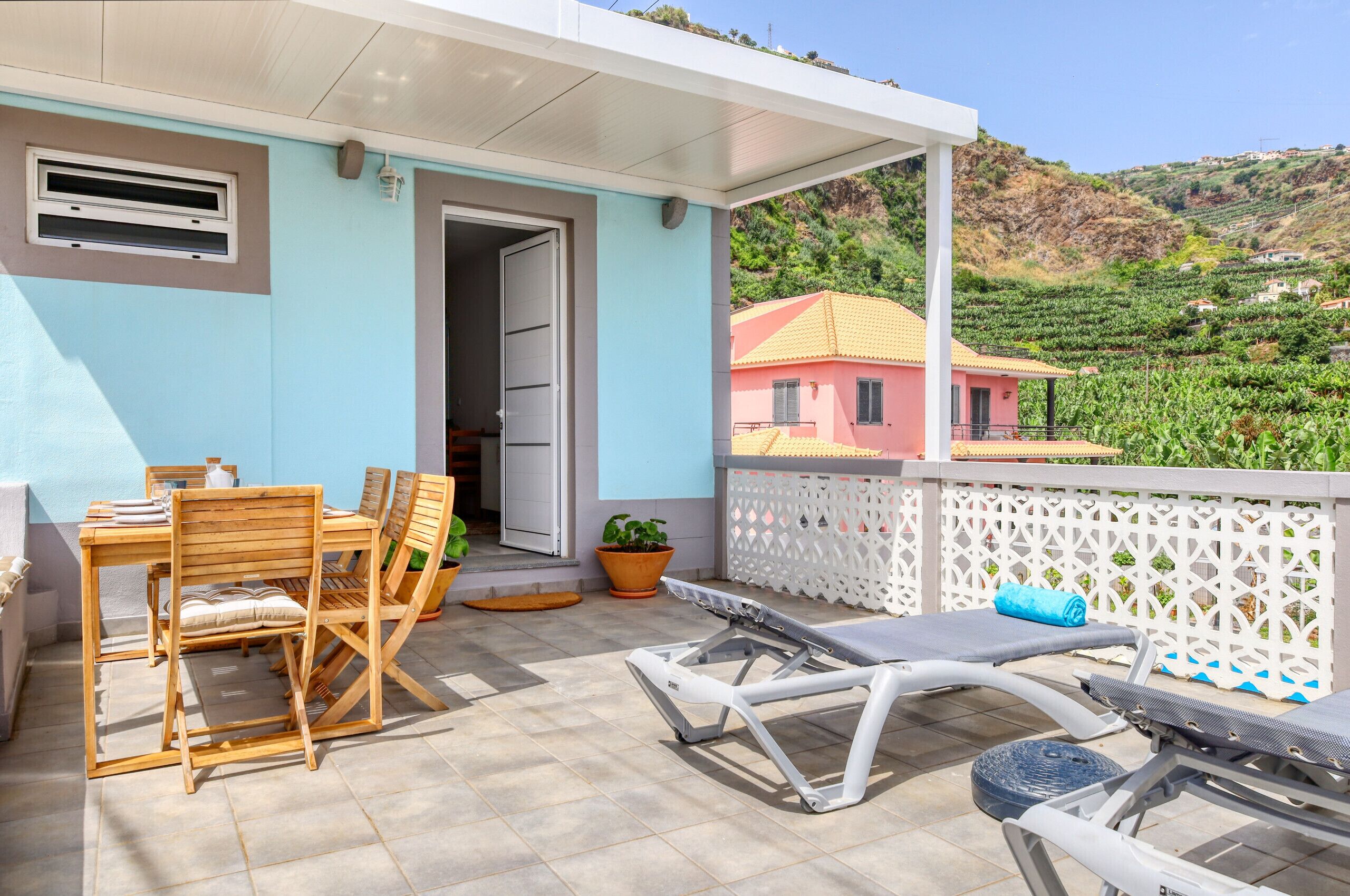 A covered outdoor area with a table and chairs, a small garden, and a view of a hillside with buildings.