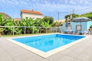 Une piscine rectangulaire bordée de blanc, entourée d’une terrasse carrelée. On y trouve des chaises longues avec des coussins bleus et un parasol gris. Un bâtiment avec un toit recouvert de tuiles rouges est visible à l’arrière-plan.