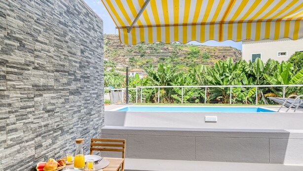 A dining area with a table set for breakfast, a pool, and a view of a hillside with lush greenery. - Aunt Celeste's House III, a Home in Madeira (Ponta do Sol)