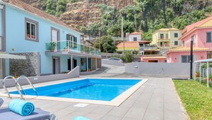 A swimming pool with lounge chairs and a blue towel, surrounded by houses with balconies and a mountainous backdrop. - Aunt Celeste's House III, a Home in Madeira (Ponta do Sol)