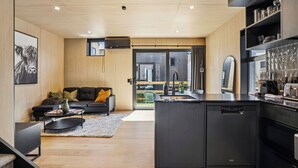 Una cocina moderna con lavavajillas integrado, un comedor con sofá negro y una pequeña mesa de centro, y una sala de estar con sofá negro y un amplio ventanal.