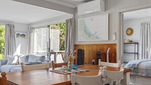 A spacious living area with a wooden dining table, white chairs, a sofa, and a bed. A map is displayed on the wall, and there is a window with curtains.