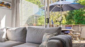 A grey sofa with a checkered throw blanket, a window with blinds, and an outdoor patio area with a table and chairs.