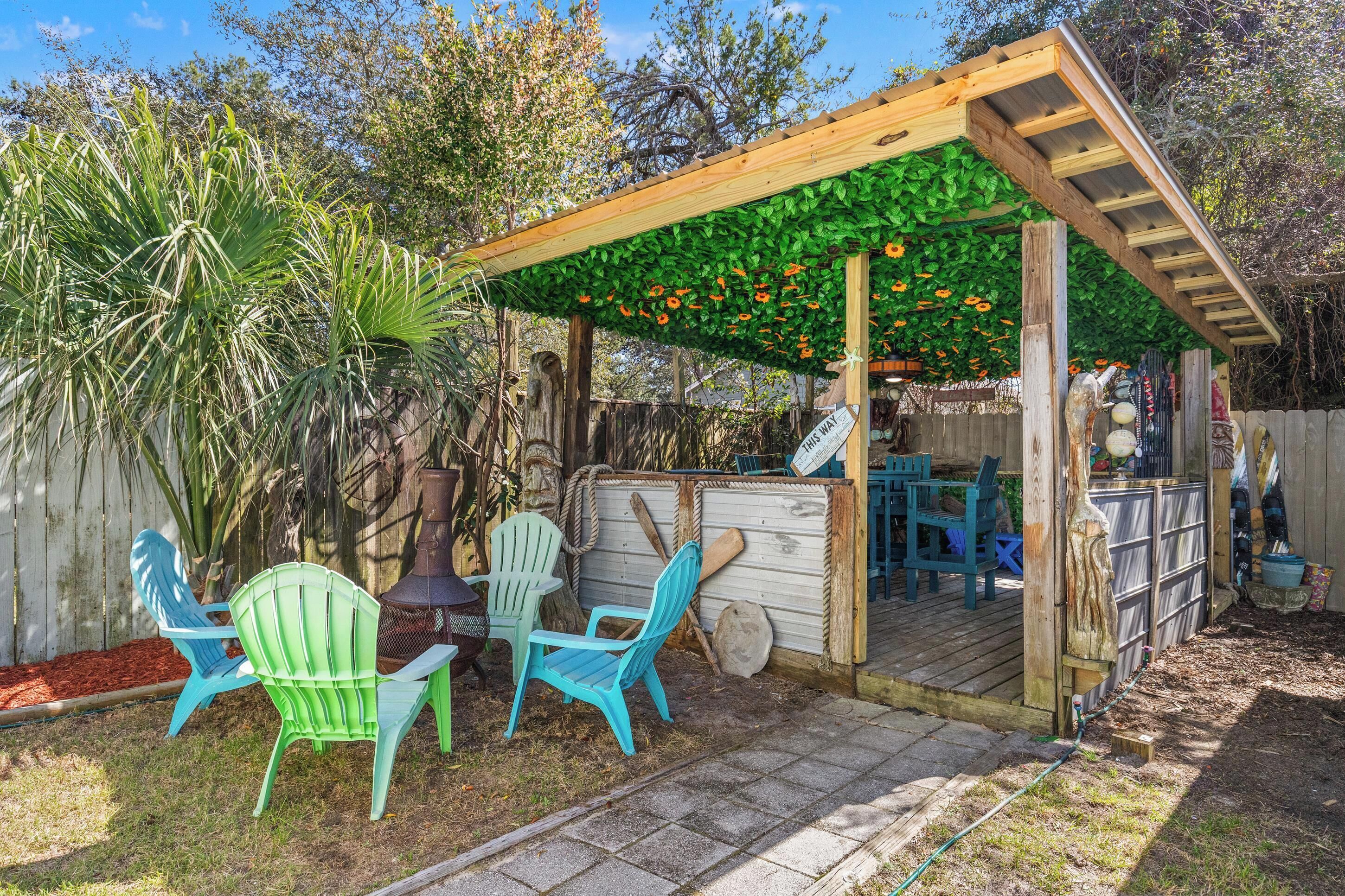Una pérgola de madera con techo verde, mobiliario exterior con sillas y una terraza de madera.