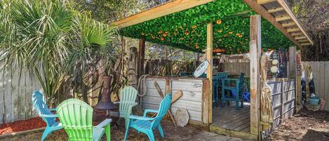 Una pérgola de madera con techo verde, mobiliario exterior con sillas y una terraza de madera.