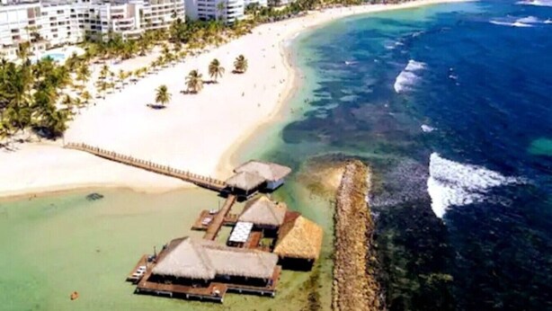 An aerial view of a beach resort with overwater bungalows, a sandy beach, and a clear blue sea.