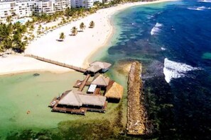 Vista aérea de un complejo turístico en la playa con bungalows sobre el agua, una playa de arena y un mar azul cristalino.