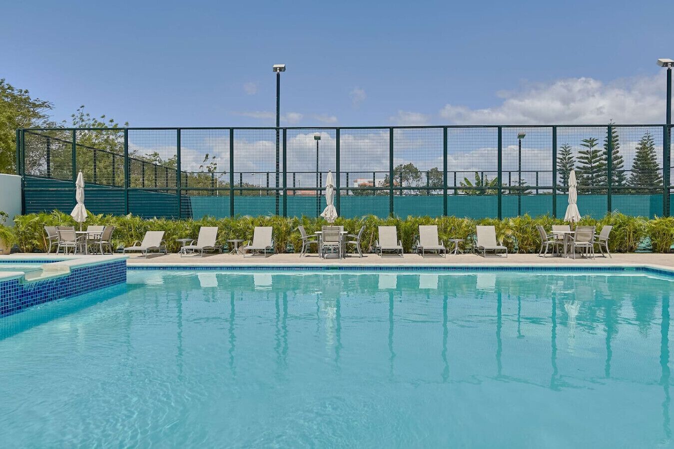 A swimming pool with lounge chairs and umbrellas, surrounded by a fence and trees.
