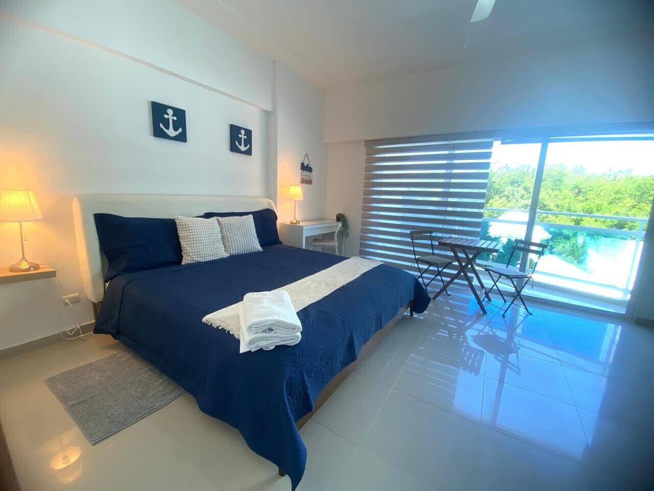 A bedroom with a large bed, two bedside lamps, a view of greenery through the window, and a small table with chairs.