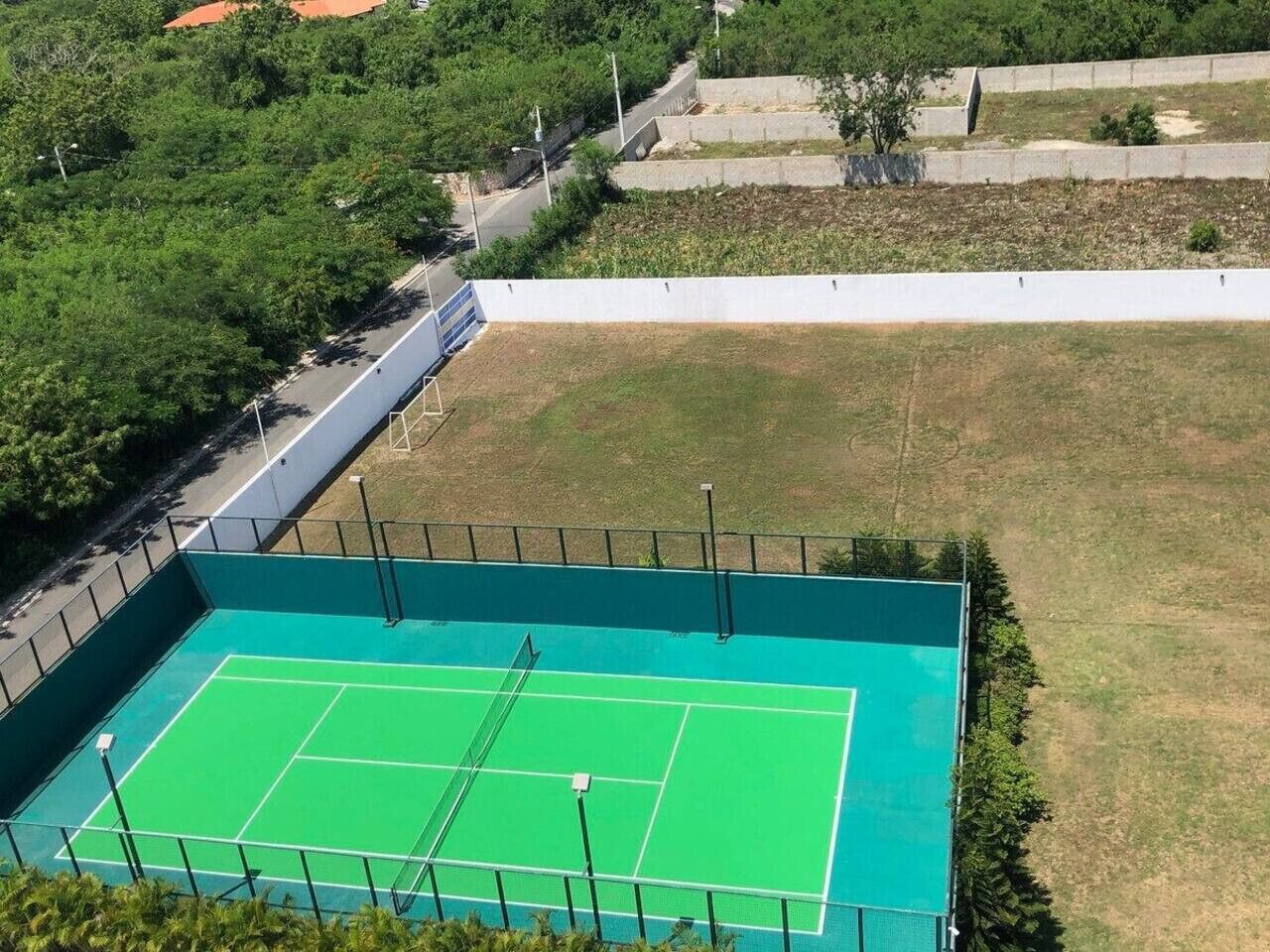 Un court de tennis entouré d’une clôture, avec des arbres et une route à proximité.