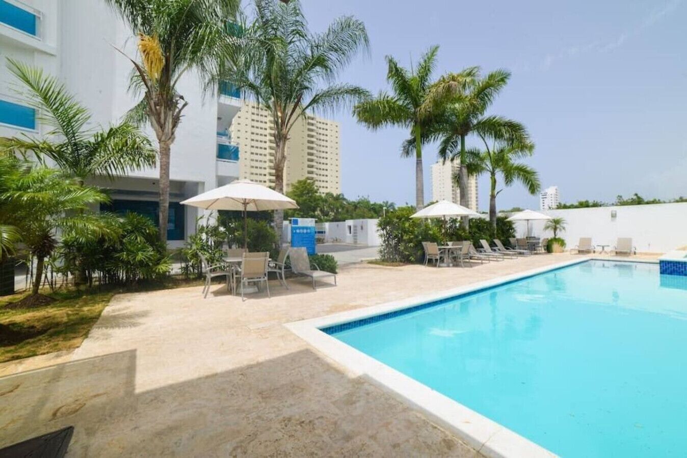 Una piscina con sillas de descanso y sombrillas, rodeada de palmeras y edificios modernos.