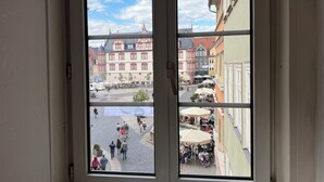 Vista a través de una ventana que muestra una calle con edificios, una fuente y terrazas.