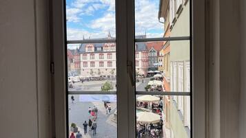 Blick durch ein Fenster auf eine Straße mit Gebäuden, einem Brunnen und Außenbereichsbestuhlung.