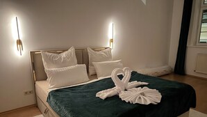 Una cama bien hecha con cubrecama verde y almohadas blancas, dos lámparas de pared y una escultura de toallas.